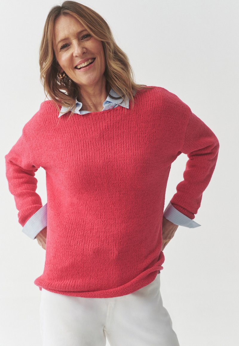Maglione rosa lavorato a maglia con una vestibilità ampia, scollatura larga e maniche lunghe. Sotto si trova una camicia azzurra con colletto. Indossato con pantaloni bianchi.