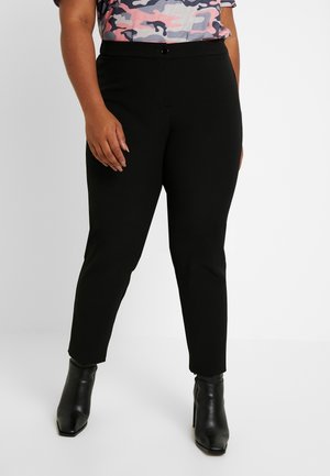 Vrouw draagt zwarte aansluitende enkelbroek, zwarte enkellaarsjes met hoge hakken en een top met camouflagepatroon in roze, grijs en wit.