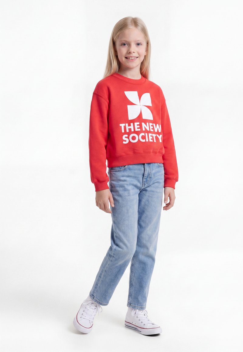Egy fiatal lány mosolyog, piros pulóvert visel fehér szöveggel „THE NEW SOCIETY“, világoskék farmert és fehér tornacipőt, fehér háttéren áll.