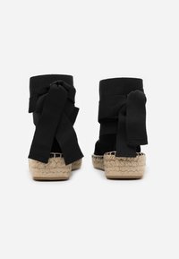 Paio di espadrillas nere alla caviglia con suole in juta intrecciata e larghe fasce in tessuto, viste da dietro.