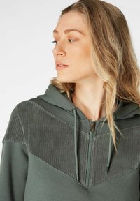 Groene hoodie met een ritssluiting aan de voorkant, voorzien van een geribbelde borstpaneel, een capuchon met trekkoord en een gladde textuur. Geen patronen of hardware details zichtbaar.