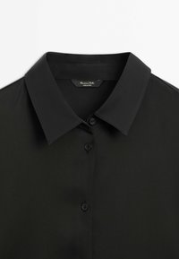 Chemise noire à boutons avec col pointu et étiquette de marque visible à l'intérieur de l'encolure, posée à plat sur fond blanc.