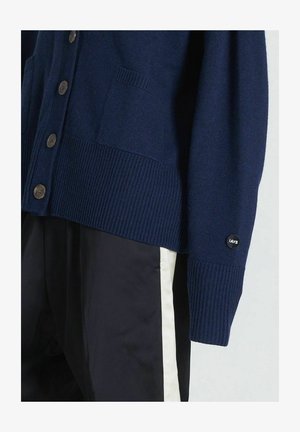 Tamsiai mėlynos spalvos megztas kardiganas su raukšlėmis ant rankogalių ir apvado, turintis priekinius kišenę ir rudus mygtukus; logotipo žymė ant rankovės.