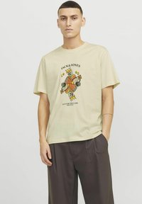 Bežinė medvilninė marškinė su ryškiu grafiniu dizainu, kuriame yra karūna, gėlės ir tekstas „JACK&JONES 1990“ juoda spalva. Trumpomis rankovėmis.