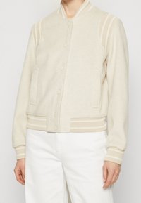 Blouson aviateur couleur crème en mélange de laine douce, avec poignets et col côtelés, fermeture à boutons-pression, et poches latérales.