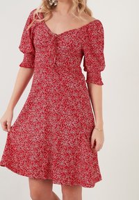 Robe fleurie rouge avec manches courtes bouffantes, encolure carrée et boutons décoratifs sur le devant. Le tissu présente de petits motifs floraux blancs.