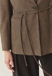 Rjav blazer s houndstooth vzorom in z pasom, ki vključuje en sam �črn gumb in teksturirano tkanino.