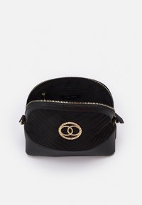 Černá prošívaná kabelka přes rameno se zipovým uzávěrem, s pozlaceným zkrouceným kruhovým akcentem vepředu a měkkou látkovou podšívkou.