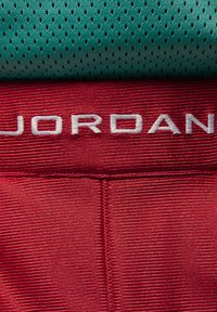 Crvene sportske šortse s teksturiranim materijalom, s bijelim "JORDAN" logotipom na pojasu, iznad zelene mrežaste majice s rupama za ventilaciju.