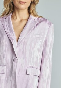 Blazer lilla caratterizzato da un tessuto testurizzato con sottili motivi lineari, rever a punta, chiusura con un solo bottone e due tasche frontali.