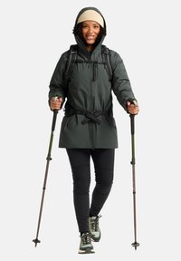 Grüne wasserdichte Jacke mit geraffter Taille, schwarze Leggings und Wanderstöcke. Verfügt über eine Kapuze und strukturierter Stoff. Passt gut zu Wanderschuhen.