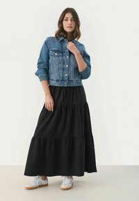 Jeansjacka med stora framfickor, knäppning med knappar och uppvullade ärmar, kombinerad med en svart lager-på-lager maxikjol och grå sneakers.