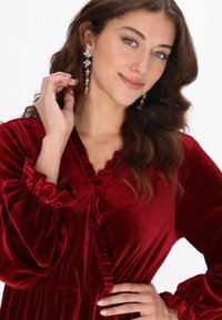 Mélyvörös bársonyruha fodros nyakkivágással és ujjakkal, alakra szabott derékkal és légiesen hullámzó szoknyával. Hosszú, lógó fülbevalók emelik ki az összhatást.