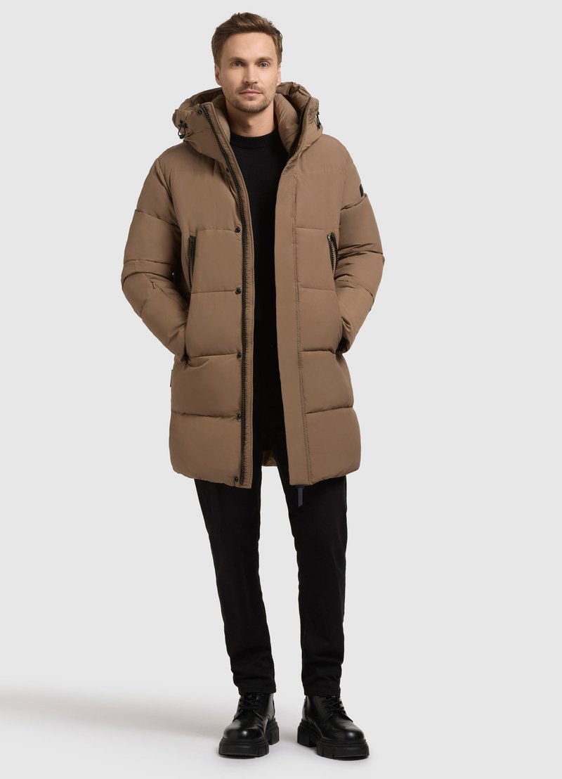 Hnedá prešívaná parka s kapucňou, ktorá má zips, postranné vrecká a prešívanú textúru, nosená cez čierny outfit a čierne topánky.