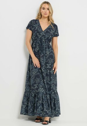 Mujer rubia con un vestido maxi azul marino con estampado floral, manga corta y dobladillo con volantes, combinado con sandalias negras con tacón.