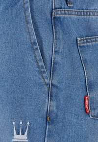 Blaue Denim-Jeans mit einer Vordertasche, kontrastierendem orangefarbenem Nähten und einem weißen Kronenlogo mit einem roten Markenschild. Strukturierten Stoff.