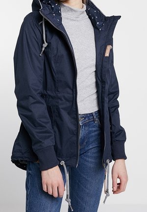 Personne portant une veste à capuche bleu marine avec cordons, un pull col roulé gris et un jean bleu, debout devant un fond uni.
