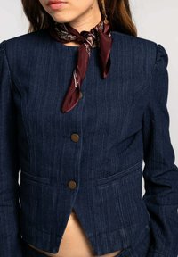Chaqueta de mezclilla en azul oscuro con rayas verticales, adornada con botones de bronce y un corte en V profundo. Combinada con una bufanda burdeos alrededor del cuello.