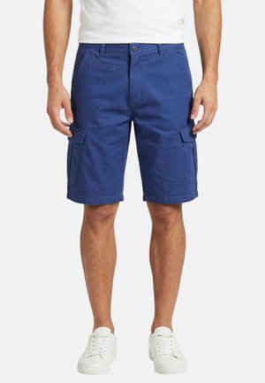 Homme portant un short cargo bleu et des baskets blanches, debout les mains détendues le long du corps, sur un fond uni.