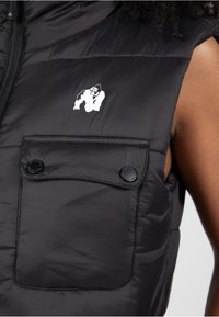 Zwarte puffer vest met een ritssluiting, twee drukknoopborstzakken en een klein wit gorilla-logo aan de linkerkant.