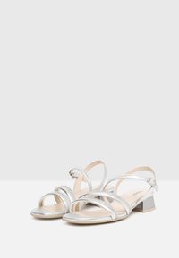 Sandales à talons hauts argentées avec des lanières croisées, finition métallique, petit talon carré et accents décoratifs en strass sur les lanières.