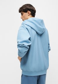 Νεαρός άνδρας με ανοιχτό μπλε φαρδιά φούτερ με κουκούλα και ταιριαστό παντελόνι, ποζάρει κοιτάζοντας στο πλάι με τα χέρια στις τσέπες, μπροστά σε απλό φόντο.