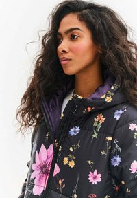 Černá prošívaná bunda s květinovým potiskem v různých barvách. Má kapuci, prošívanou texturu a výrazné růžové a fialové květy.