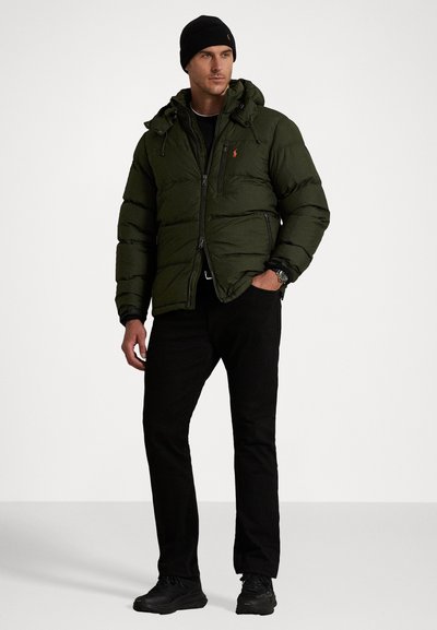 Mand iført en mørkegrøn pufferjakke, sort beanie, sorte bukser og sorte sneakers står med den ene hånd i lommen.