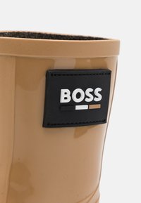 Botte en caoutchouc beige avec une finition brillante. Présente une étiquette noire avec "BOSS" en blanc, accompagnée d'accents noirs et marron. Texture lisse.