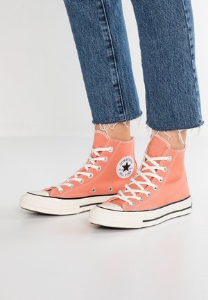 Person iført koralfarvede høje Converse All Star sneakers med hvide snørebånd og slidte blå denimjeans på en hvid gulv.