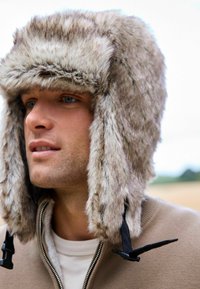 Faux-Fur-Trappermütze in Grautönen mit Ohrenklappen, die eine weiche Textur und schwarze verstellbare Riemen an den Seiten aufweist.