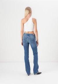 Frau mit langen blonden Haaren steht mit dem Rücken zur Kamera, trägt ein weißes One-Shoulder-Crop-Top, blaue Jeans und schwarze High Heels.