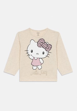 T-shirt beige à manches longues avec un grand motif Hello Kitty portant une robe rose et un nœud à pois. "Hello Kitty" écrit en paillettes en dessous.
