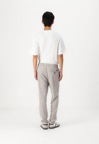 Man staat met de rug naar ons toe in een wit T-shirt met korte mouwen, lichtgrijze broek met achterzak, en witte sneakers met zwarte strepen.