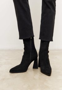 Botas de tobillo de ante negro con punta afilada y tacón grueso. Presentan un diseño limpio, una forma ajustada y una textura suave.