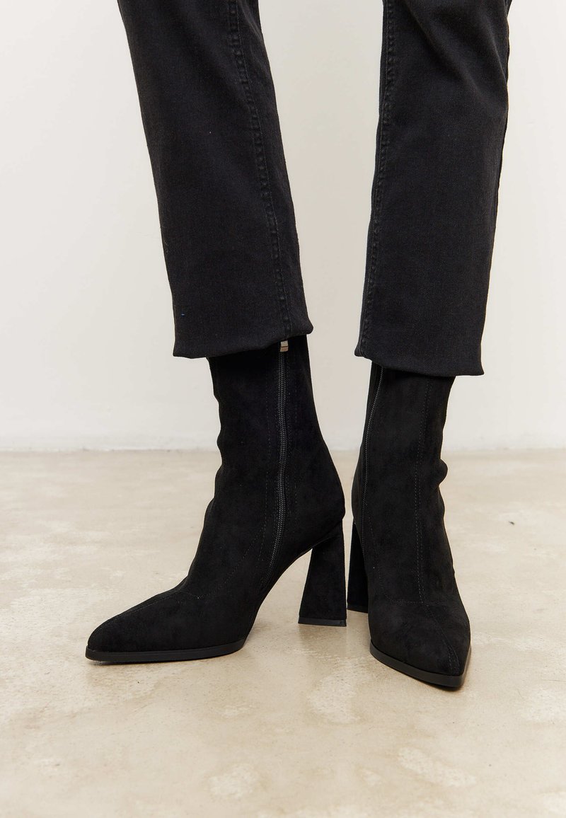 Botas de tobillo de ante negro con punta afilada y tacón grueso. Presentan un diseño limpio, una forma ajustada y una textura suave.