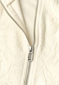 Fermeture éclair argentée partiellement ouverte sur un tissu crème texturé à motif floral, mettant l'accent sur le curseur et les dents de la fermeture.