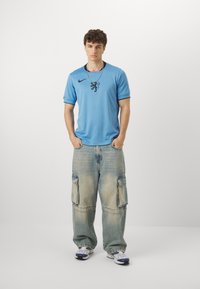 Maglietta Nike blu con logo di un leone nero, abbinata a pantaloni cargo in denim sbiadito e larghi. Il modello indossa sneakers bianche con dettagli blu navy. Collana semplice.
