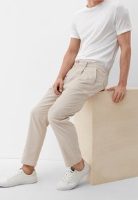 Beige broek met elastische taille en twee voorzakken, gecombineerd met een witte T-shirt met ronde hals en witte sneakers, zittend op een licht houten blok.