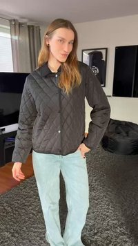 Sort quiltet jakke med krave, knaplukning og lange ærmer, kombineret med lyseblå jeans på et tekstureret tæppe.