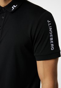 Svart polo skjorta med mjuk textur, knäppbar krage och broderad vit logotyp och varumärkesnamn på kragen och ärmen.