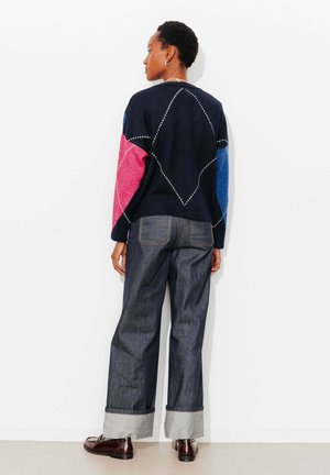 Personne debout de dos portant un pull marine avec des manches roses et bleues, un jean à jambes larges retroussé et des chaussures marron foncé.