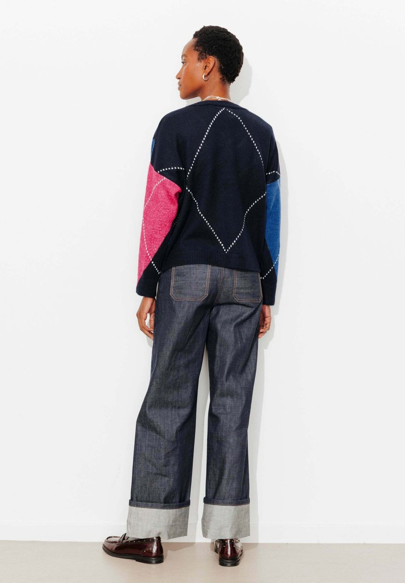 Personne debout de dos portant un pull marine avec des manches roses et bleues, un jean à jambes larges retroussé et des chaussures marron foncé.