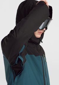 Chaqueta de esquí con capucha y mangas negras, cuerpo color teal, con cremalleras y un tejido texturizado. Incluye gafas de nieve llevadas en la cabeza.