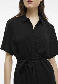 Juodas, trumpomis rankovėmis marškinėlių suknelė iš tekstūruoto audinio, su apykakle, mygtukų uždarymu priekyje ir surišamu juosmeniu. Nėra matomų raštų ar akcentų.