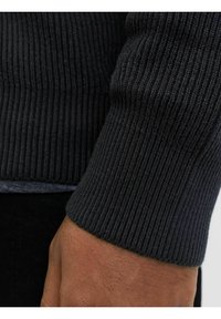 Rebrasti crni džemper rukav prikazuje teksturirani pleteni uzorak, s glatkim rubom i vidljivim šavom. Ruka tamnije kože naslonjena je na rukav.