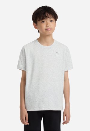 Garçon aux cheveux courts et foncés portant un t-shirt gris clair à manches courtes avec un petit logo de cerf en haut à gauche et un pantalon noir.