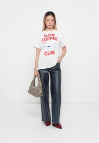 Sieviete, tērpusies baltā T-kreklā ar uzrakstu "Slow Coffee Club", tumšos platās biksēs, sarkanos apavos, tur rokās čūskādaino somu, stāv uz balta fona.