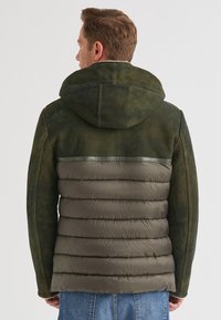 Mann in grün-grauer, gepolsterter Jacke mit Kapuze, der von hinten vor einem schlichten Hintergrund steht.