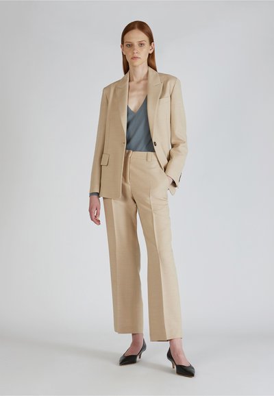 Beige Anzug mit weitem Hosenbein und einem Blazer mit einem Knopf, kombiniert mit einem blauen V-Ausschnitt-Oberteil und schwarzen Absatzschuhen.